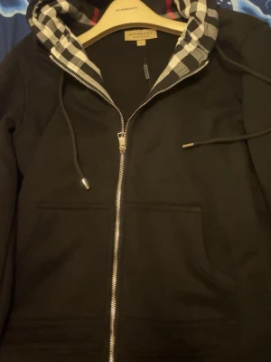 Svart hoodie från Burberry med dragkedja - Svart hoodie från Burberry med klassisk dragkedja framtill och två fickor. Huvan har det ikoniska rutmönstret i svart, vitt och grått samt dragsko. Perfekt för en stilren och lyxig streetlook.