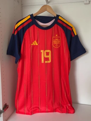 Spaniens landslagströja Lamine Yamal - Röd och gul fotbollströja från Adidas för Spaniens landslag med nummer 19 och namnet Lamine Yamal på ryggen. Tröjan har blå ärmar, gula ränder på axlarna och tunna vertikala linjer framtill. Perfekt för dig som älskar fotboll och vill stötta Spanien.