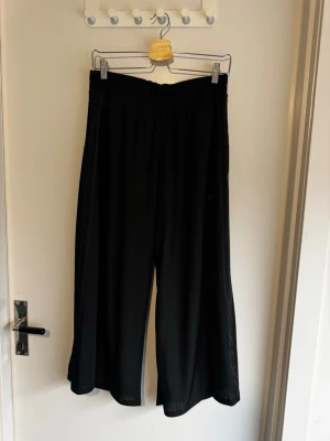 Svarta vida croppade mjukisbyxor från Adidas - Svarta croppade mjukisbyxor från Adidas med vida ben och diskret broderad logga på benet. Byxorna har resår i midjan och är gjorda i ett lätt, plisserat material som ger en snygg och avslappnad look.