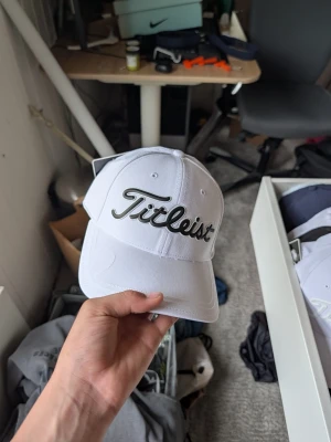 Vit Titleist keps med broderad logga - Snygg vit keps från Titleist med broderad svart logga framtill. Klassisk böjd skärm och justerbar rem baktill för perfekt passform. Tillverkad i ett lätt och ventilerande material som passar perfekt för golf eller vardagsstil.