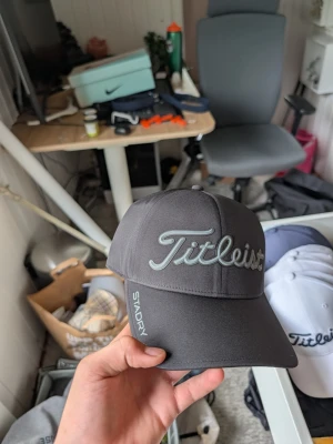 Mörkgrå Titleist keps med brodyr - Snygg mörkgrå keps från Titleist med broderad logga framtill och texten 'STADRY' på sidan. Klassisk böjd skärm och justerbar baksida. Perfekt accessoar för golf eller streetwear. Materialet är troligtvis polyester för vattenavvisande effekt.