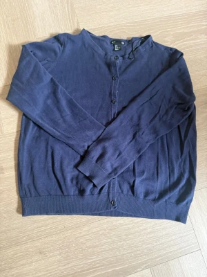 Mörkblå cardigan från Lindex - Mörkblå cardigan från Lindex med rund halsringning och knäppning framtill. Långärmad modell i mjukt, tunt material med ribbade muddar vid ärmslut och nederkant. Perfekt att slänga över en t-shirt eller linne.