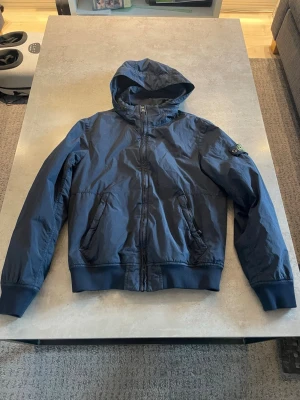 Mörkblå vindjacka från Stone Island - Snygg mörkblå vindjacka från Stone Island med huva och dragkedja framtill. Jackan har två fickor med dragkedja och klassisk Stone Island-logga på vänster ärm. Perfekt för blåsiga dagar och streetwear-stil.