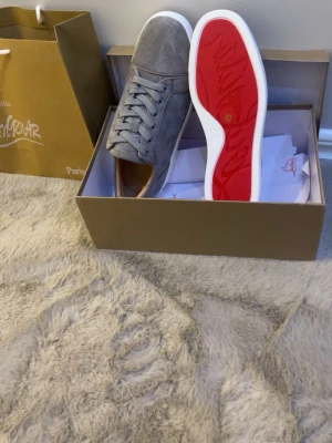 Gråa Christian Louboutin sneakers - Snygga gråa sneakers från Christian Louboutin i mocka med klassisk röd sula och vita kanter. Skorna har rund tå, gråa skosnören och diskret branding. Perfekta för dig som vill ha en stilren och lyxig look.