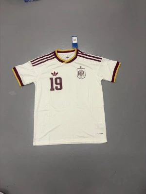 Spaniens Vm tröja Lamine - Säljer en vit Spanien fotbollströja från Adidas med nummer 19 och Lamine Yamal på ryggen. Tröjan har vinröda och gula detaljer, korta ärmar och klassiska Adidas-ränder på axlarna. Perfekt för dig som älskar fotboll och vill stötta landslaget. Storlek S