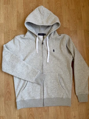 Grå zip hoodie från Polo Ralph Lauren - Snygg ljusgrå hoodie med dragkedja från Polo Ralph Lauren. Klassisk design med vit dragsko i huvan, två fickor fram och den ikoniska mörkblå loggan broderad på bröstet. Tillverkad i mjuk bomullsblandning som är perfekt för chill dagar. Den är otroligt skön och helt ny och oanvänd. Skriv vid frågor eller funderingar.