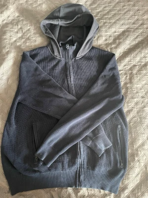 Mörkgrå zip-hoodie med fickor - En mörkgrå hoodie med dragkedja framtill och två fickor med dragkedja. Huvan är i mjukare tyg än resten av tröjan, som har en stickad struktur. Perfekt för en chill och avslappnad stil.