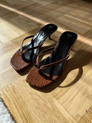 Bruna sandaler med klack från Zara - Säljer ett par trendiga bruna sandaler från Zara med fyrkantig tå och smala remmar. Sandalerna har låg klack och croco-mönstrad yta i konstläder. Använt ett fåtal gånger.