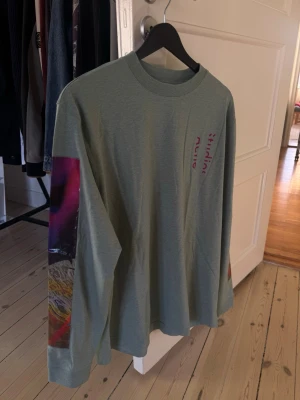 Grönblå långärmad t-shirt Acne Studios - Väldigt unik runway piece från Acne. Köpt på Acne Archive. Snygg grönblå långärmad t-shirt från Acne Studios med tryck ärmarna och diskret logga på bröstet. Lätt oversized passform och rund halsringning. Perfekt för dig som gillar streetwear och unika detaljer. Storlek S men passar M/L