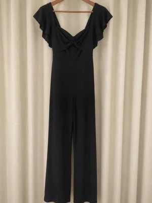 ASOS Svart jumpsuit med volangärm - Stilren svart jumpsuit med vida ben och djup v-ringning. Volangärmar ger en trendig touch och twistad detalj framtill. Tillverkad i ett mjukt, följsamt tyg som sitter snyggt på kroppen. Perfekt för dig som gillar en modern och elegant look.