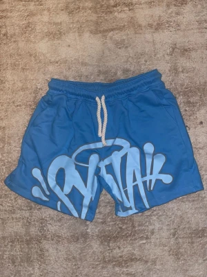 Blå shorts med graffiti-tryck - Blå shorts med vitt graffiti-inspirerat tryck framtill och vit snörning i midjan. Shortsen har elastisk midja och en bakficka med dragkedja. Perfekta för sommaren och har en avslappnad passform.