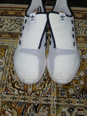 Vita Adidas slip-on sneakers - Snygga vita slip-on sneakers från Adidas med svart kant och detaljer. Ovandelen är i meshmaterial som andas och har en modern, minimalistisk look utan snörning. Perfekta för dig som gillar stilrena och bekväma skor med sportig känsla.