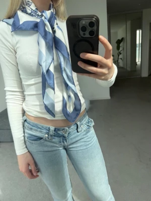 Blåvit mönstrad sidenscarf - Snygg scarf i siden med blått och vitt mönster. Kan stylas både runt halsen och som midjesjal, eller på väska!. Scarfen har en glansig finish och klassiskt paisleymönster blandat med ränder. Perfekt accessoar för att lyfta din outfit.