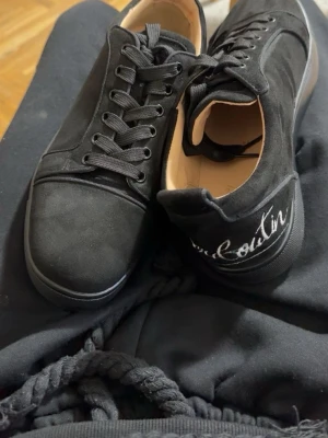 Svarta sneakers från Christian Louboutin - Snygga svarta sneakers från Christian Louboutin i mocka med klassisk låg siluett. Skorna har svarta skosnören, diskret sömdetalj och broderad logga på hälen. Perfekta för dig som gillar stilrena och exklusiva sneakers.