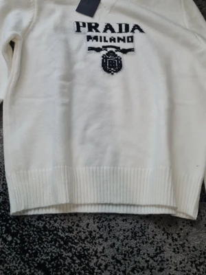 Vit stickad tröja från Prada - Vit stickad tröja från Prada med svart broderad logotyp och texten 'PRADA MILANO' framtill. Tröjan har ribbstickade muddar vid ärmslut och nederkant. Klassisk och stilren design som passar perfekt till många outfits.