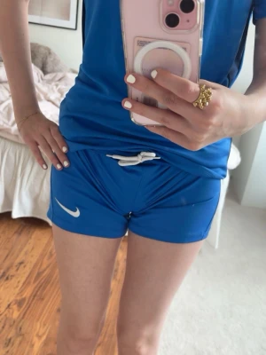 träningsshorts - Snygga blå shorts från Nike med vit logga på benet och praktisk snörning i midjan. Perfekta för träning eller sportiga dagar. De har en klassisk passform och är gjorda i ett lätt och bekvämt material.