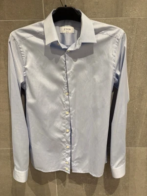 Ljusblå skjorta från Eton  - Snygg ljusblå skjorta från Eton i modellen Super Slim. Skjortan har klassisk krage, långa ärmar och vita knappar. Perfekt för dig som gillar en stilren och slimmad look. Tillverkad i ett tunt och mjukt tyg som känns lyxigt mot huden.