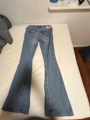 True Religion Joey Low Rise Flare Jeans - Säljer ett par ljusblå True Religion Joey jeans med låg midja och utsvängda ben. Klassiska bakfickor med lock och snygga sömmar.