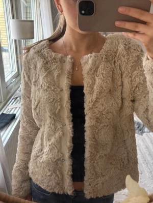 Beige teddyjacka med knappar - Mysig beige teddyjacka från vero moda i storlek Xs. Jackan har en rund halsringning och knäppning framtill. Jackan har en fluffig och mjuk struktur som ger en trendig look. Perfekt för att hålla värmen under kyliga dagar. Passar till jeans eller kjol för en avslappnad stil.