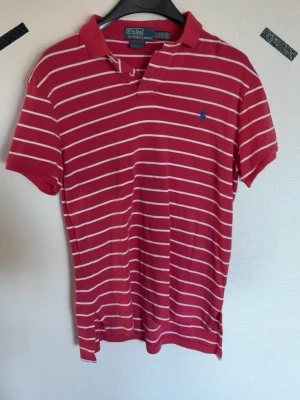 Röd- och vitrandig pikétröja Polo Ralph Lauren - Röd pikétröja från Polo Ralph Lauren med vita horisontella ränder och klassisk krage. Tröjan har korta ärmar och blå broderad logga på bröstet. Perfekt för en avslappnad och stilren look.