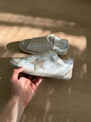 Golden Goose vita sneakers med stjärna - Snygga vita sneakers från Golden Goose med beige mockadetaljer, stor stjärna på sidan och glittrig silverdetalj vid hälen. Skorna har klassisk låg profil och vita snören. Perfekta för dig som gillar en unik och trendig look. 💕💕💕