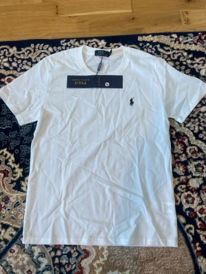 Vit t-shirt från Polo Ralph Lauren - Klassisk vit t-shirt från Polo Ralph Lauren med rund halsringning och ikonisk broderad logga på bröstet. Tillverkad i mjuk bomull och har en normal passform. Perfekt basplagg för en stilren look.