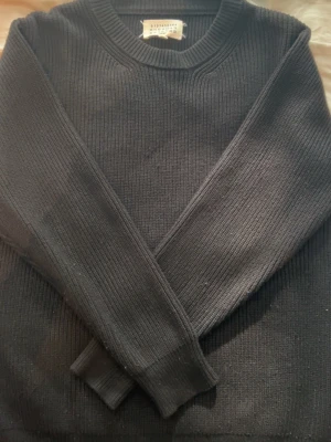 Svart stickad tröja från maison margiela - En stilren svart stickad tröja från maison margiela med rund halsringning och ribbade muddar. Tröjan har en klassisk passform och är tillverkad i ett mjukt, värmande material som passar perfekt till kyligare dagar.