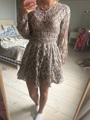 Blommig kortklänning med lång ärm - Supersöt kortklänning med blommigt mönster i beige, brunt och lila. Klänningen har långärmade ärmar, markerad midja med smock och lätt vippig kjol. Tillverkad i ett tunt och luftigt material, perfekt för vår och sommar.