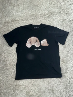 Palm Angels svart t-shirt med björn - Svart t-shirt från Palm Angels med ikoniskt björntryck på framsidan och stor logga i vitt på ryggen. Klassisk rund hals och korta ärmar. Tillverkad i mjuk bomull för skön känsla och snygg streetwear-look. Storlek: S/M