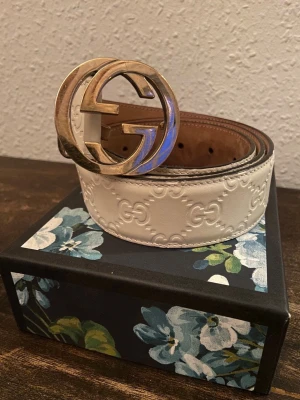 Vit skinnbälte med GG-spänne från Gucci - Säljer ett exklusivt vitt skinnbälte från Gucci med präglat GG-mönster och ett stort, glänsande guld GG-spänne. Bältet är tillverkat i Italien och har en lyxig känsla som verkligen sticker ut. Perfekt för att lyfta din outfit.