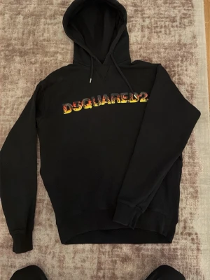 Dsquared2 hoodie - Snygg svart hoodie från Dsquared2 med färgglad logga i rött, gult och orange på bröstet. Klassisk huva med dragsko och ribbade muddar vid ärmar och nederkant. Perfekt för en trendig och avslappnad stil.