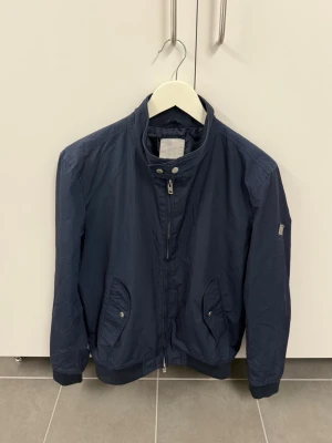 Marinblå vindjacka från J.Lindeberg stl M - Snygg marinblå vindjacka från J.Lindeberg med hög krage och dragkedja framtill. Jackan har två fickor med tryckknappar och ett diskret logomärke på ärmen. Materialet är lätt och passar perfekt för blåsiga dagar.
