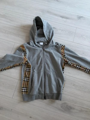 Grå hoodie med rutiga detaljer - Säljer en grå hoodie från burberry med dragkedja och huva. Hoodien har snygga rutiga detaljer i beige, svart och rött längs ärmarna och sidorna. Materialet är mjukt och bekvämt, perfekt för chill dagar. Stora fickor framtill och ribbade muddar.