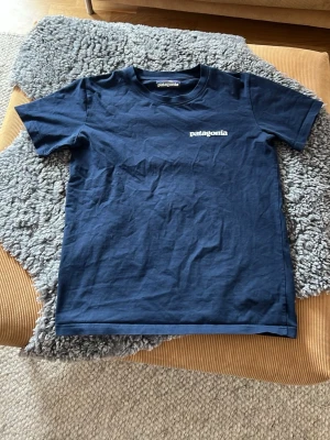 Marinblå Patagonia t-shirt - Säljer en marinblå t-shirt från Patagonia med klassisk logga på bröstet och ett stort färgglatt tryck med berg och logotyp på ryggen. T-shirten har korta ärmar och är gjord i mjuk bomull. Perfekt för dig som gillar outdoor-vibes och enkel stil.