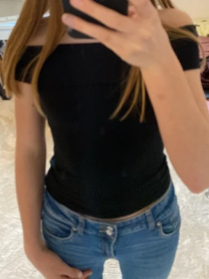 Svart offshoulder topp - Snygg svart offshoulder topp med tight passform. Toppen är ärmlös och har en bred kant över axlarna som ger en trendig look. Perfekt att matcha med jeans eller kjol för en stilren outfit..