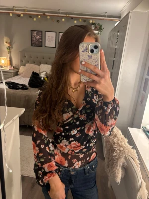 Blommig mesh body  - Säljer en snygg body från newyorker i storlek XS. Den är gjord i transparent mesh-material med svart bas och stora rosor i orange och vitt, samt gröna blad. Långärmad med djup v-ringning och knäppning i grenen. 