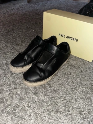 Svarta Axel Arigato sneakers - Snygga svarta sneakers från Axel Arigato i slät läderdesign med diskret guldfärgad logga på sidan. Skorna har en chunky vit sula och minimalistisk look utan synliga snören. Perfekta för dig som gillar stilrena och trendiga skor. OBS skorna är välanvända, sömmarna runt skon har släppt lite se bild.( går och fixa enligt skräddare) Vid köp har du godkänt dessa defekter och varan går inte och returnera. Hälarna är lagade en gång.