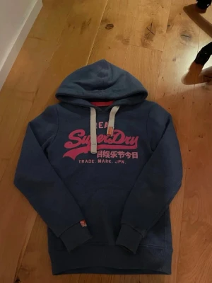 Blå Superdry hoodie med tryck - Snygg blå hoodie från Superdry med stor rosa logga och japansk text på bröstet. Tröjan har huva med vita dragsnören och ribbade muddar vid ärmar och nederkant. Perfekt för en avslappnad streetwear-look. Modellen är 163cm.