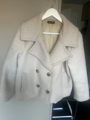 Krämvit teddy-cardigan från Chelsea - Supermjuk och fluffig krämvit cardiganjacka från Chelsea med bred krage och dubbelknäppning framtill. Jackan har två fickor och är fodrad på insidan. Perfekt för att hålla sig varm och snygg under kyliga dagar.nypris 999