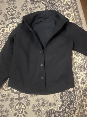  overshirt med knappar - Stilren svart overshirt med klassisk krage och knäppning framtill. Overshirten har en bröstficka och raka ärmar. Perfekt att bära över en t-shirt för en avslappnad look. Materialet ser ut att vara mjukt och något tjockare, troligen bomull eller ullblandning. Storlek XS men sitter S/M