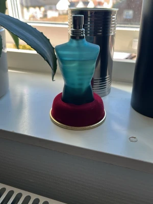 Jean Paul Gaultier Le Male parfym - Säljer en ikonisk parfym från Jean Paul Gaultier, Le Male. Flaskan är formad som en blå manlig torso med matt och transparent finish, och har en silverfärgad spraykork. Parfymen står på en rund röd sammetsbas med guldkant. De är en 75 ml flaska men de är ca 55 ml kvar