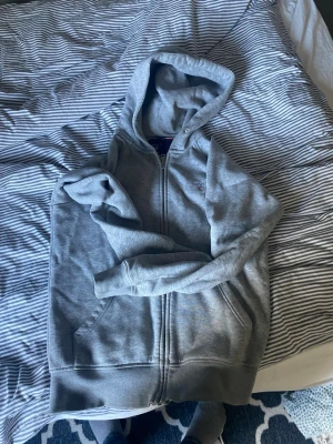 Grå zip-hoodie från Gant - Säljer en grå hoodie med dragkedja från Gant. Den har huva, två fickor framtill och diskret logga på bröstet. Perfekt för chill dagar och enkel att matcha med jeans eller joggers.