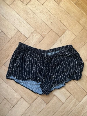 Svarta prickiga shorts med snörning - Säljer ett par svarta shorts från H&M med vita prickiga ränder och snörning i midjan. De är loose fit och har en mjuk, tunn bomullskänsla som passar perfekt till varma dagar. Elastisk midja och avslappnad stil. Strl M-L.