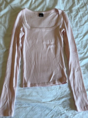 Ljusrosa ribbad långärmad topp - Säljer en ljusrosa ribbad topp med lång ärm och bred, fyrkantig halsringning. Materialet är mjukt och stretchigt, perfekt för att skapa en snygg och enkel look. Passformen är tight och toppen är enkel att matcha med jeans eller kjol.