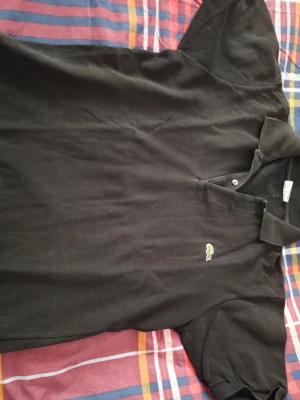 Svart Lacoste pike  - Säljer en svart pike från Lacoste med klassisk krage och två knappar framtill. T-shirten har korta ärmar och den ikoniska gröna krokodilen broderad på bröstet. Perfekt för dig som gillar stilrena plagg med en sportig touch.