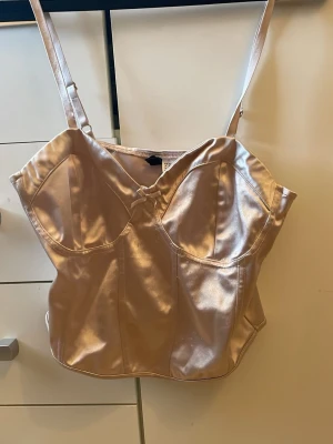 Glansig beige satintopp med rosett - Snygg beige topp i glansigt satinmaterial med smala justerbara axelband och en liten rosett framtill. Formade kupor och markerade sömmar ger en feminin siluett. Perfekt för dig som vill ha något stilrent och elegant.