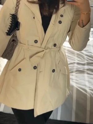 Beige trenchcoat med knytskärp - Stilren beige trenchcoat med knytskärp i midjan. Klassisk krage och två framfickor.