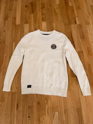 Stickat Morris tröja  - Vit stickad tröja från morris. Tröjan har ett broderat märke på bröstet och en liten svart patch vid nederkanten. Perfekt för dig som gillar stilrena plagg med diskreta detaljer.