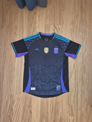  OG Retro 2022 VM Argentina away kit fotbollströja Adidas svart/lila - Säljer en svart Argentina fotbollströja från Adidas med lila och turkosa detaljer. Tröjan har korta ärmar, mönstrad framsida och logga samt mästerskapsmärke på bröstet. Perfekt för dig som vill sticka ut på planen eller heja på laget.