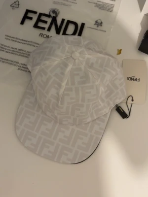 Vit Fendi keps med FF-mönster - Säljer en stilren vit keps från Fendi med det klassiska FF-monogrammet över hela kepsen. Kepsen har böjd skärm och justerbar baksida. Tillverkad i ett lätt material som passar perfekt till streetwear och trendiga outfits.
